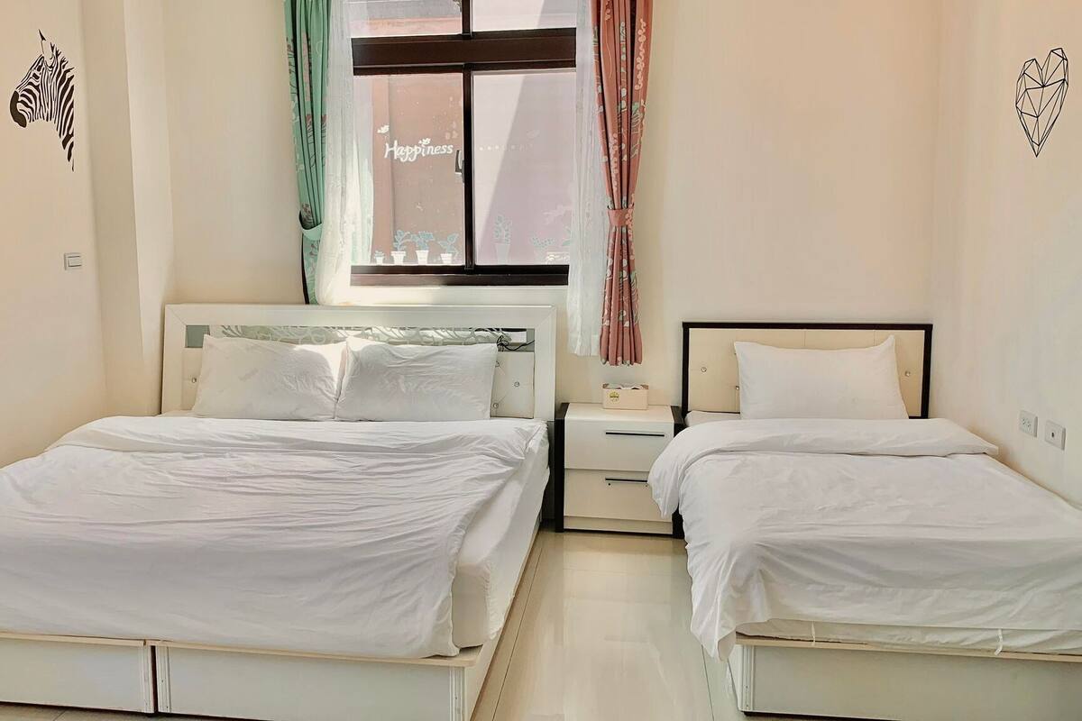 A hotel room with two single beds, a nightstand, a window with curtains, and a wall decal.