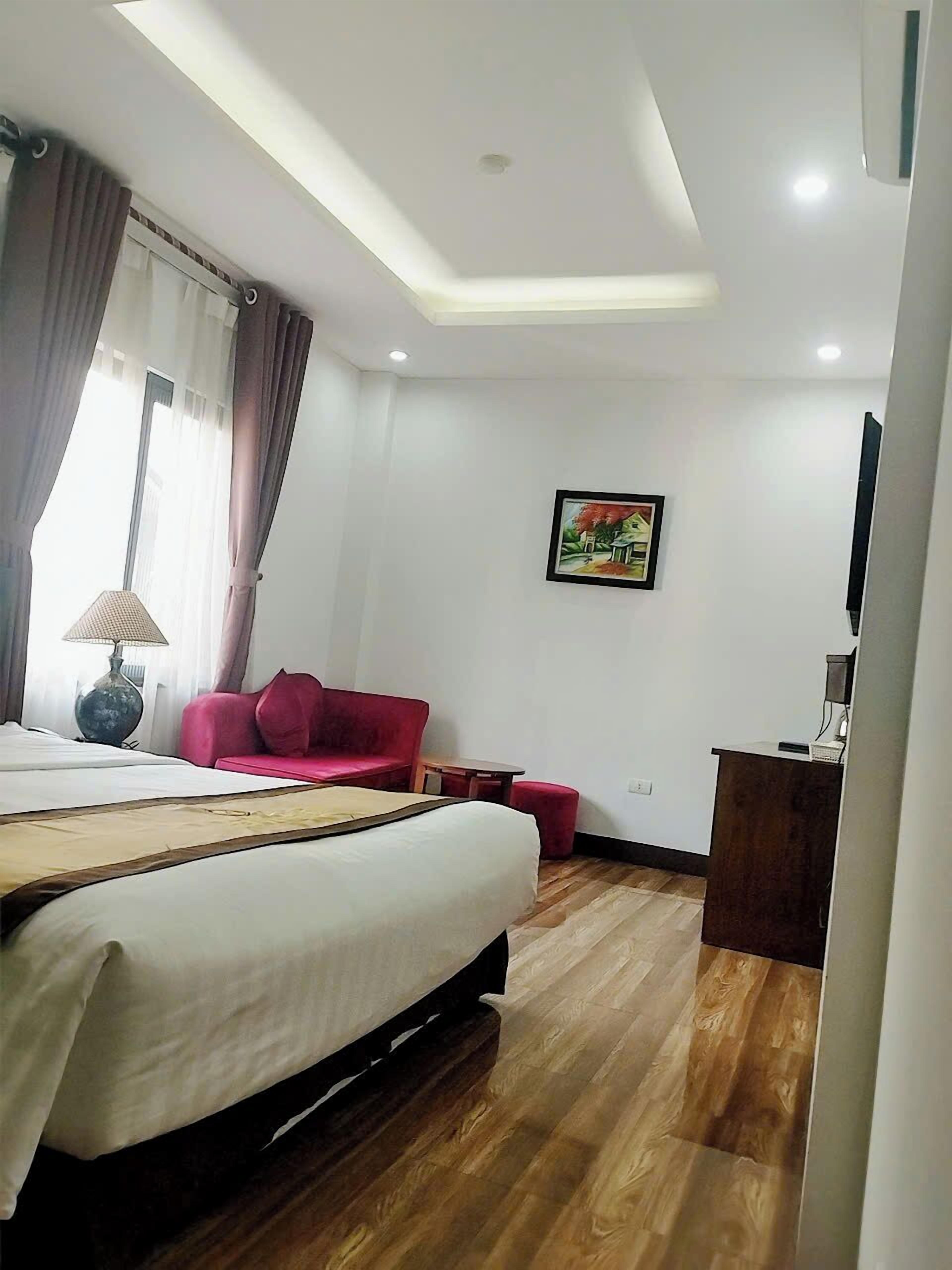 A hotel room with a bed, a red sofa, a small table, a desk, a lamp, a painting, and a ceiling light.