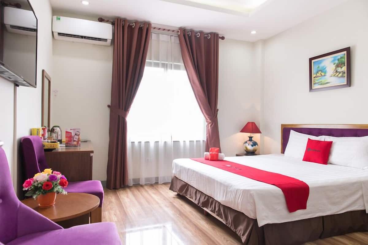 Habitación de hotel con cama, sillón púrpura, mesita con flores y un cuadro en la pared.