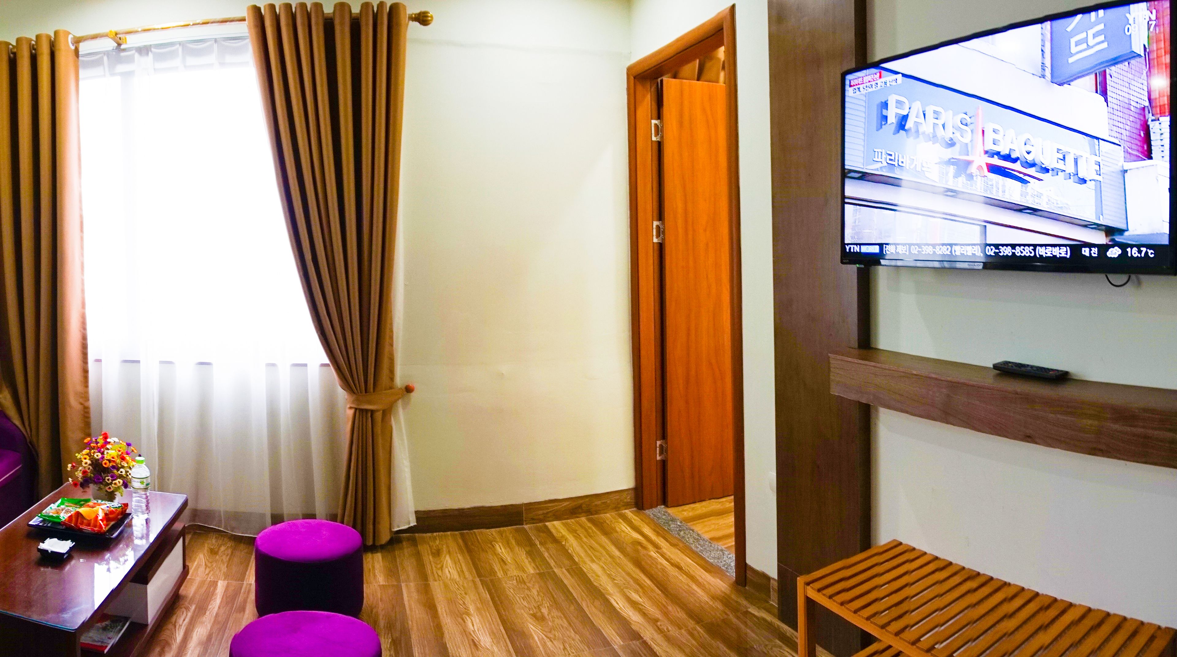Ein Hotelzimmer mit Holzboden, einem an der Wand befestigten Fernseher, einer Holztür und einem Fenster mit Vorhängen.