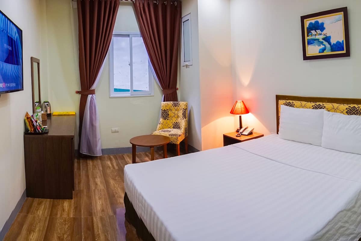 Una habitación de hotel con una cama, una silla, un escritorio con lámpara, un televisor y un cuadro en la pared.