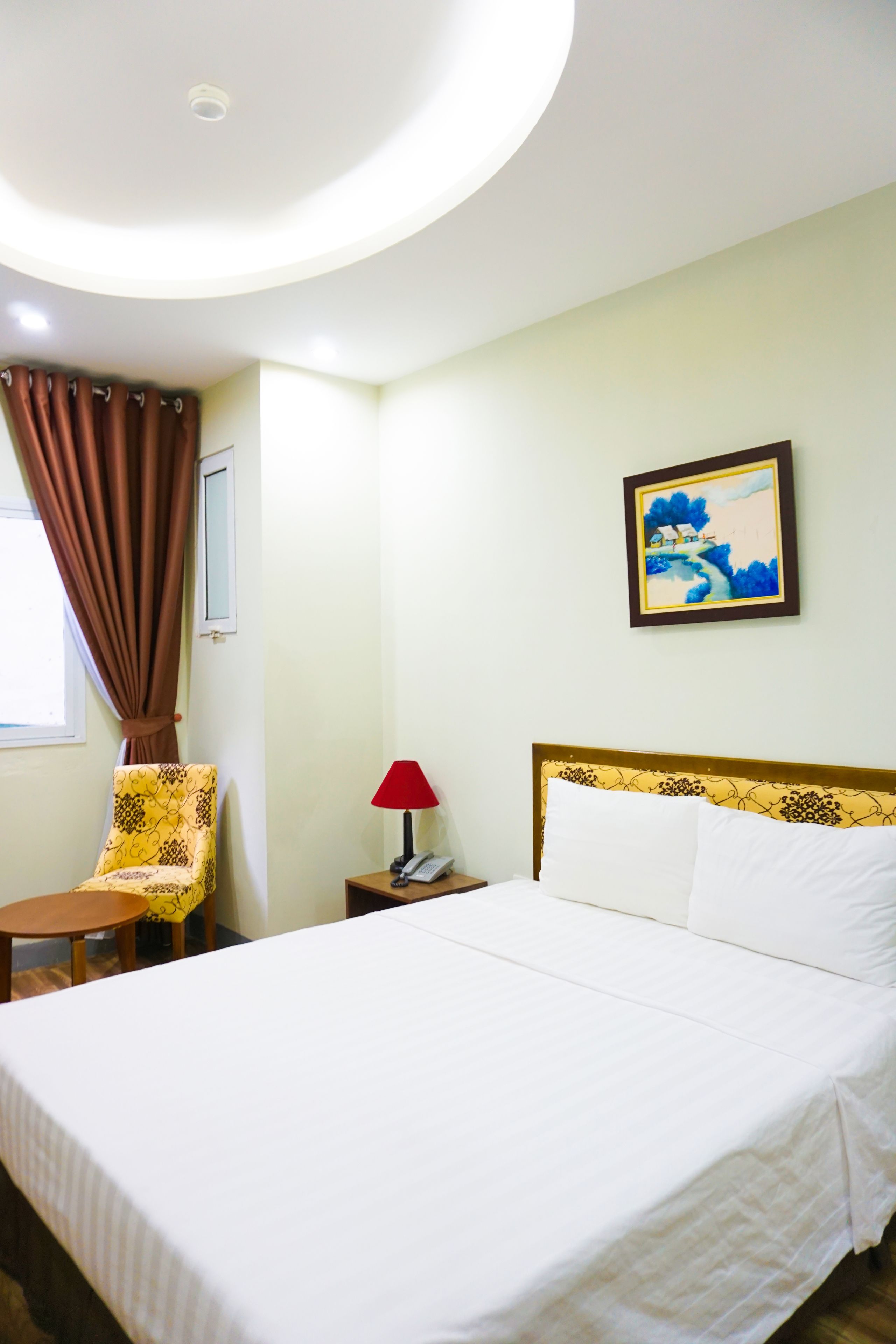 A hotel room with a bed, a chair, a nightstand, a lamp, a painting, and a window with curtains.