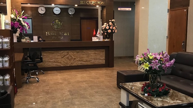 A hotel reception area with a reception desk, a clock, and a sign indicating the hotel name. There are flower arrangements on the desk and a sofa in the foreground. - K Business 3 Hotel (Formerly Mai Hotel) (Hanoi)