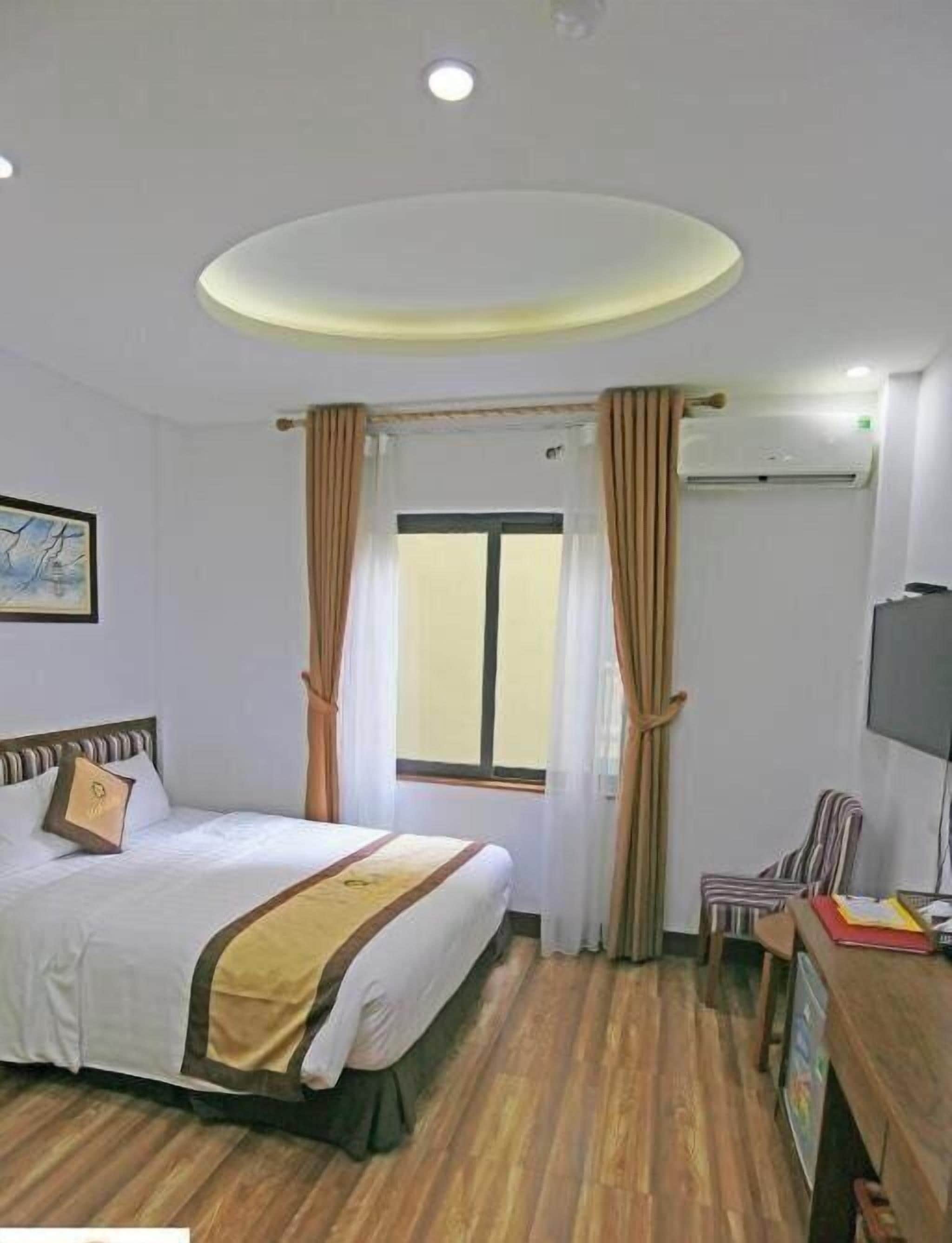 Une chambre d’hôtel comprenant un lit, un bureau, une chaise, une fenêtre avec des rideaux, un appareil de climatisation et un luminaire au plafond.