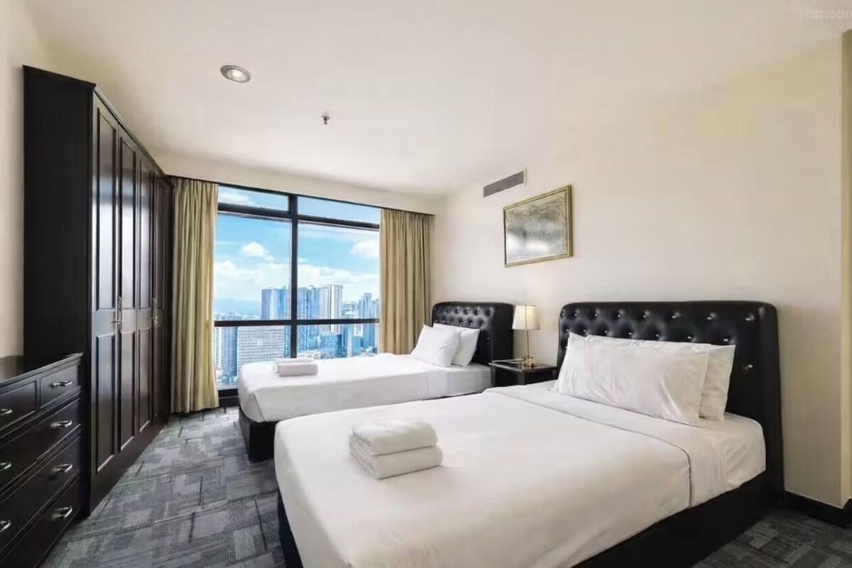 A hotel room with two beds, a wardrobe, a large window with a city view, and a framed picture on the wall.