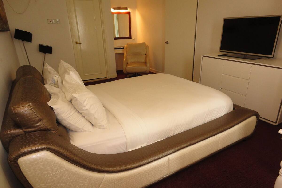 A bed with a headboard, a television on a stand, and a chair in a room with a door and a mirror.