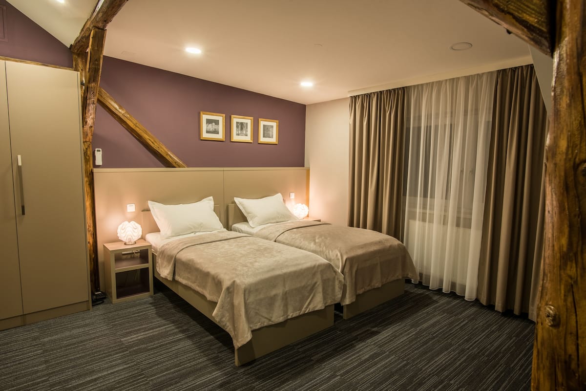 A double bed with two pillows, a nightstand with a lamp, a wardrobe, and a large window with curtains.