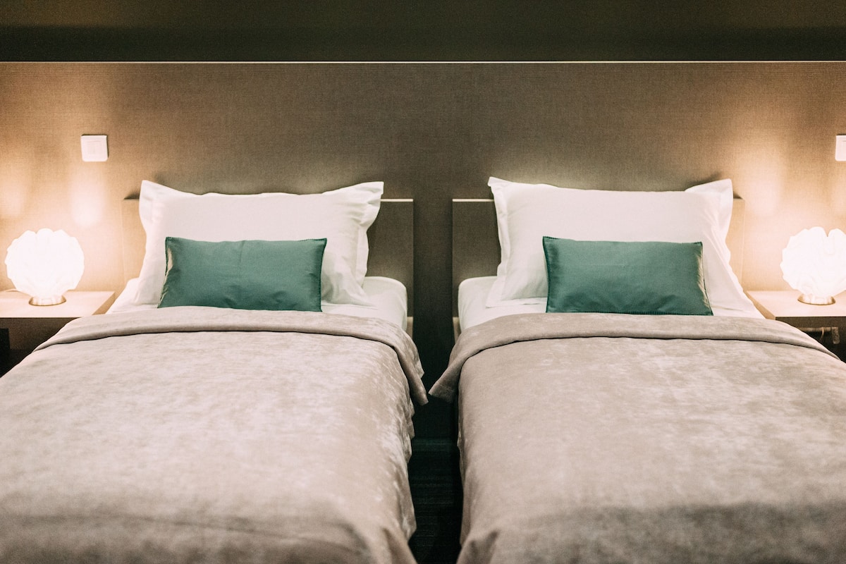 A twin room with two beds, each with a white pillow and a green pillow, and a nightstand with a lamp.
