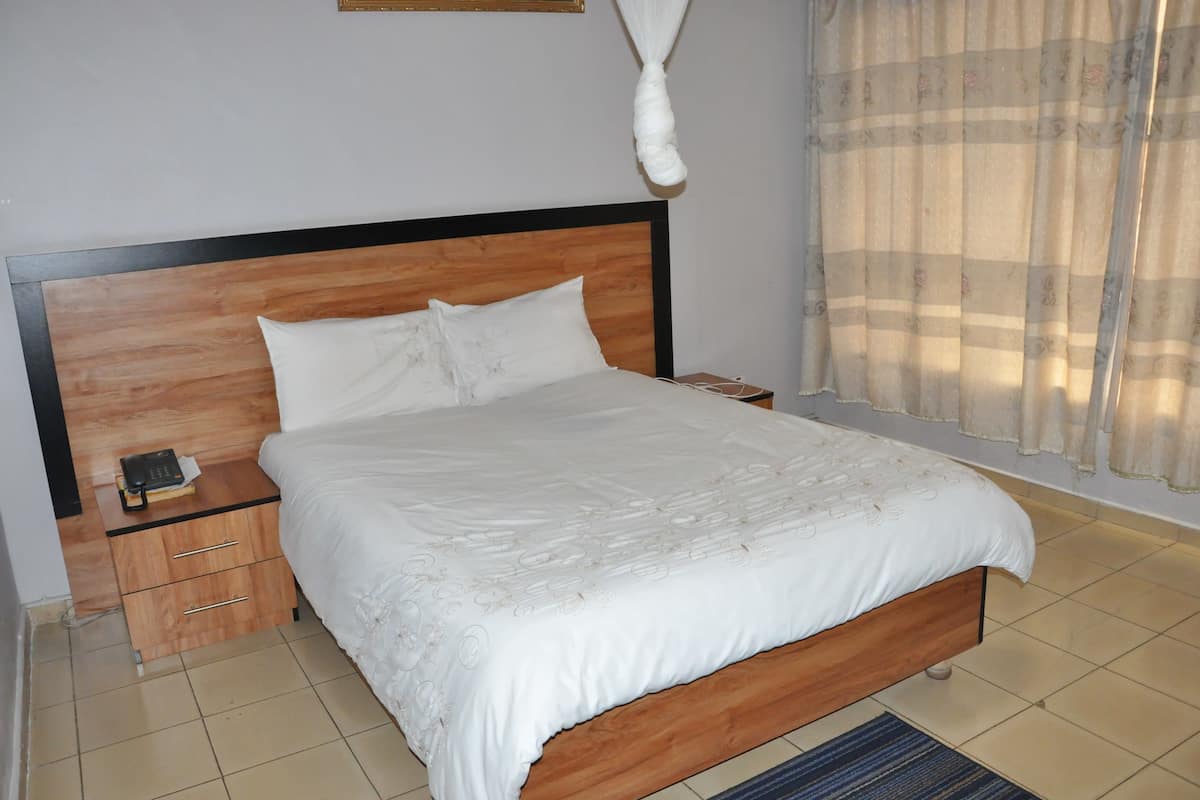 A neatly made bed with a wooden headboard, a bedside table with a phone, and a window with sheer curtains.