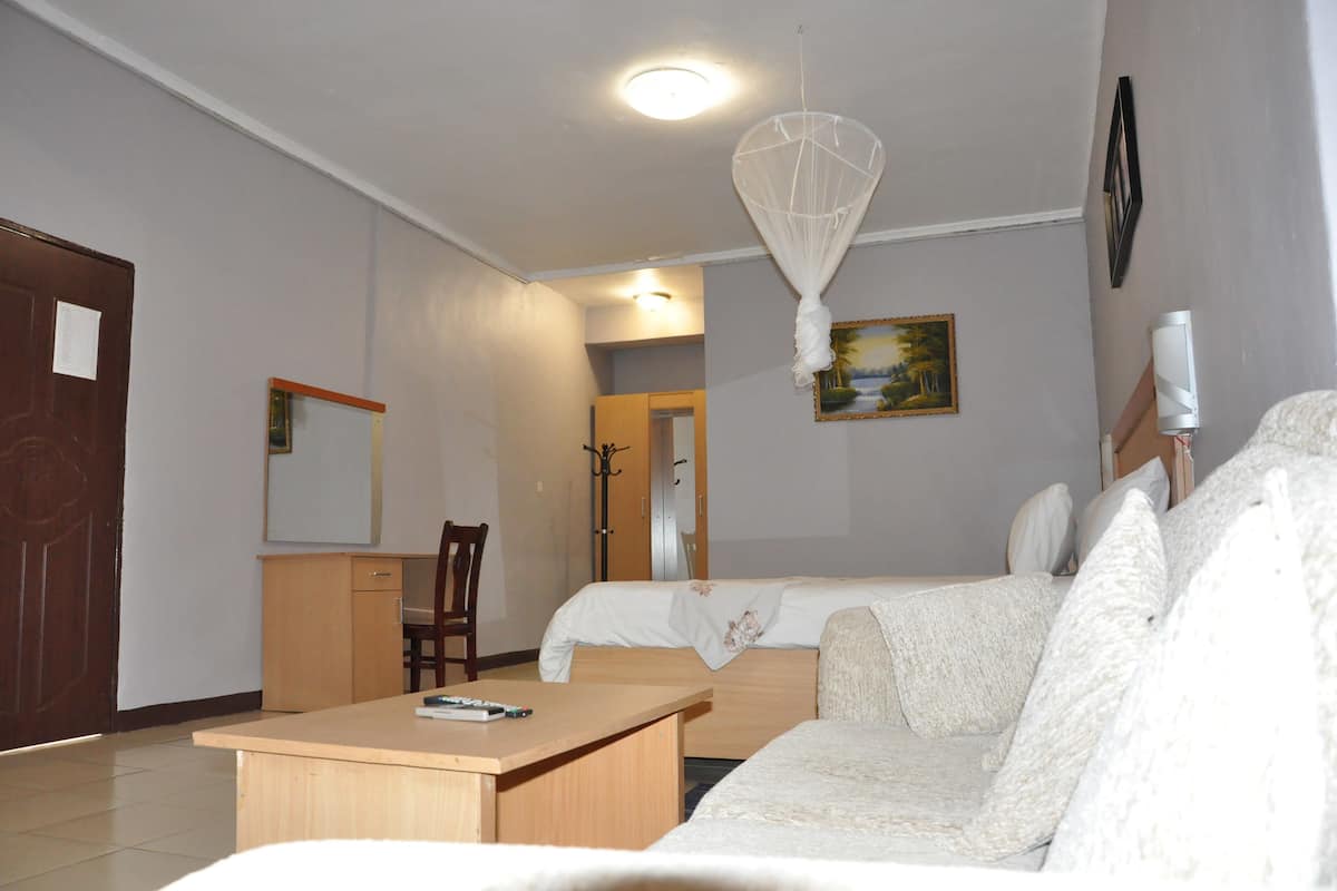 A hotel room with a bed, a desk, a chair, a small table, a hanging light fixture, and a painting on the wall.