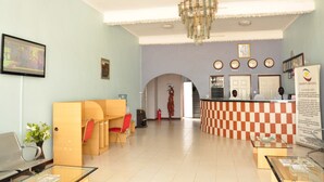 A hotel lobby with a reception desk, a chandelier, a checkered counter, and a seating area with a vase of flowers.
