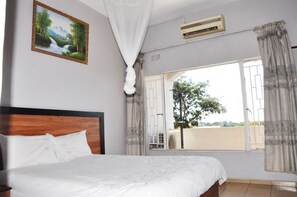 A bedroom with a bed, a window with curtains, an air conditioning unit, and a painting on the wall. - Crown Hotel Lilongwe (Lilongwe)