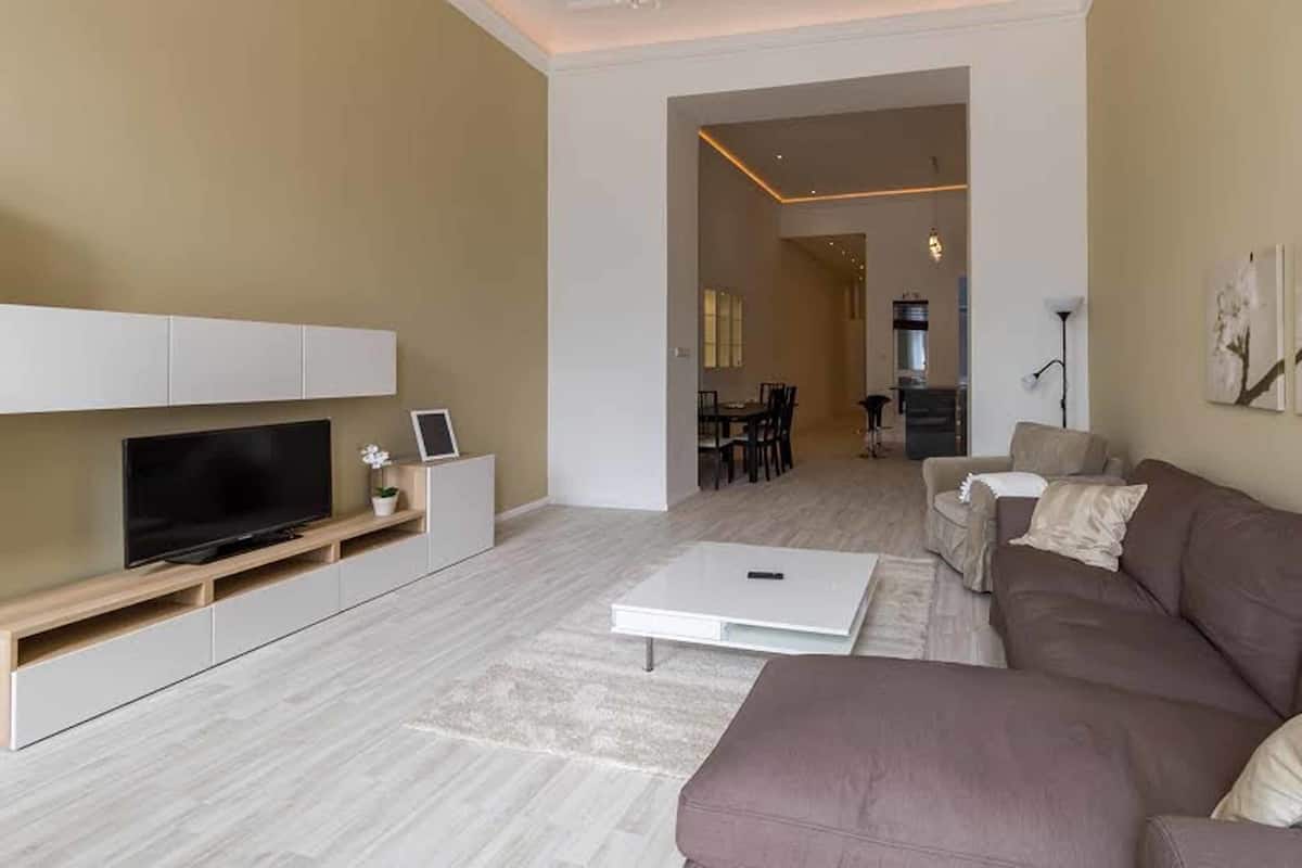 Uma sala de estar moderna com uma televisão de tela plana, um sofá, uma mesa de centro e uma área de jantar ao fundo.