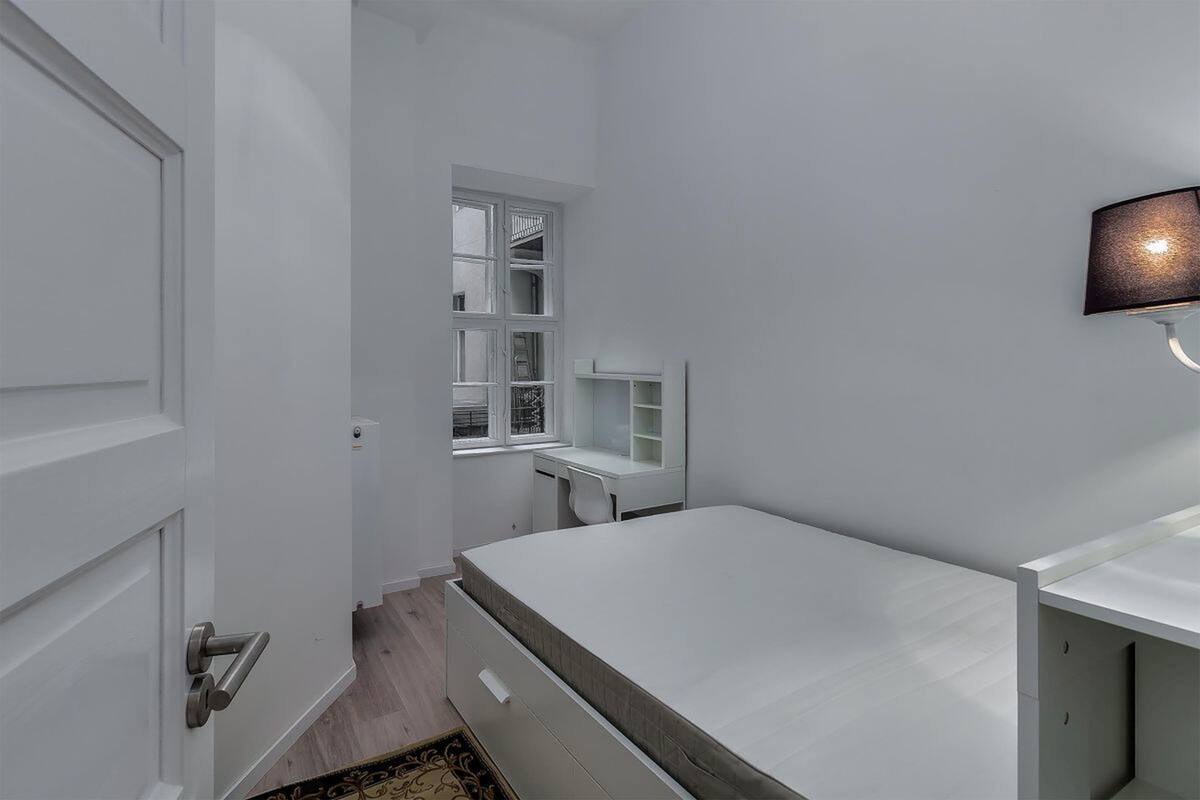 Um quarto pequeno e branco, com uma cama, uma escrivaninha e uma janela.