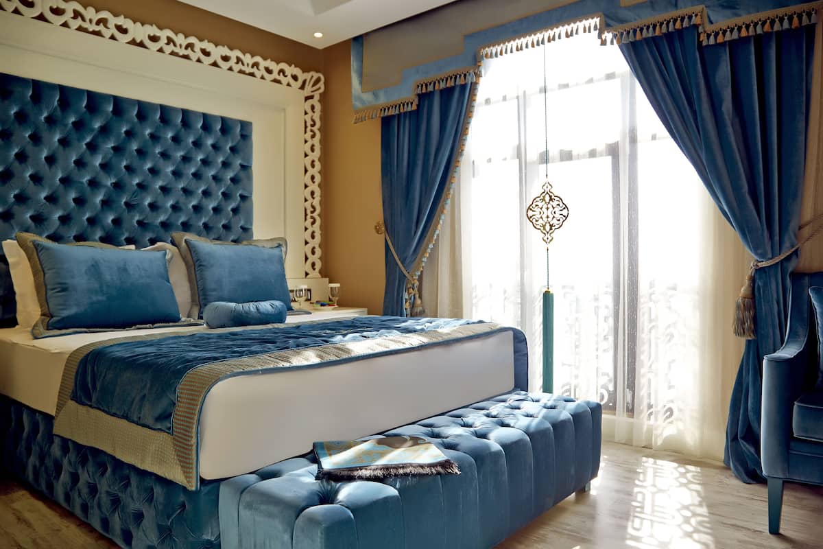 Un dormitorio con cabecera capitonada, una cama grande con ropa de cama azul, un otoman a juego y vistas del exterior a través de cortinas de gasa.