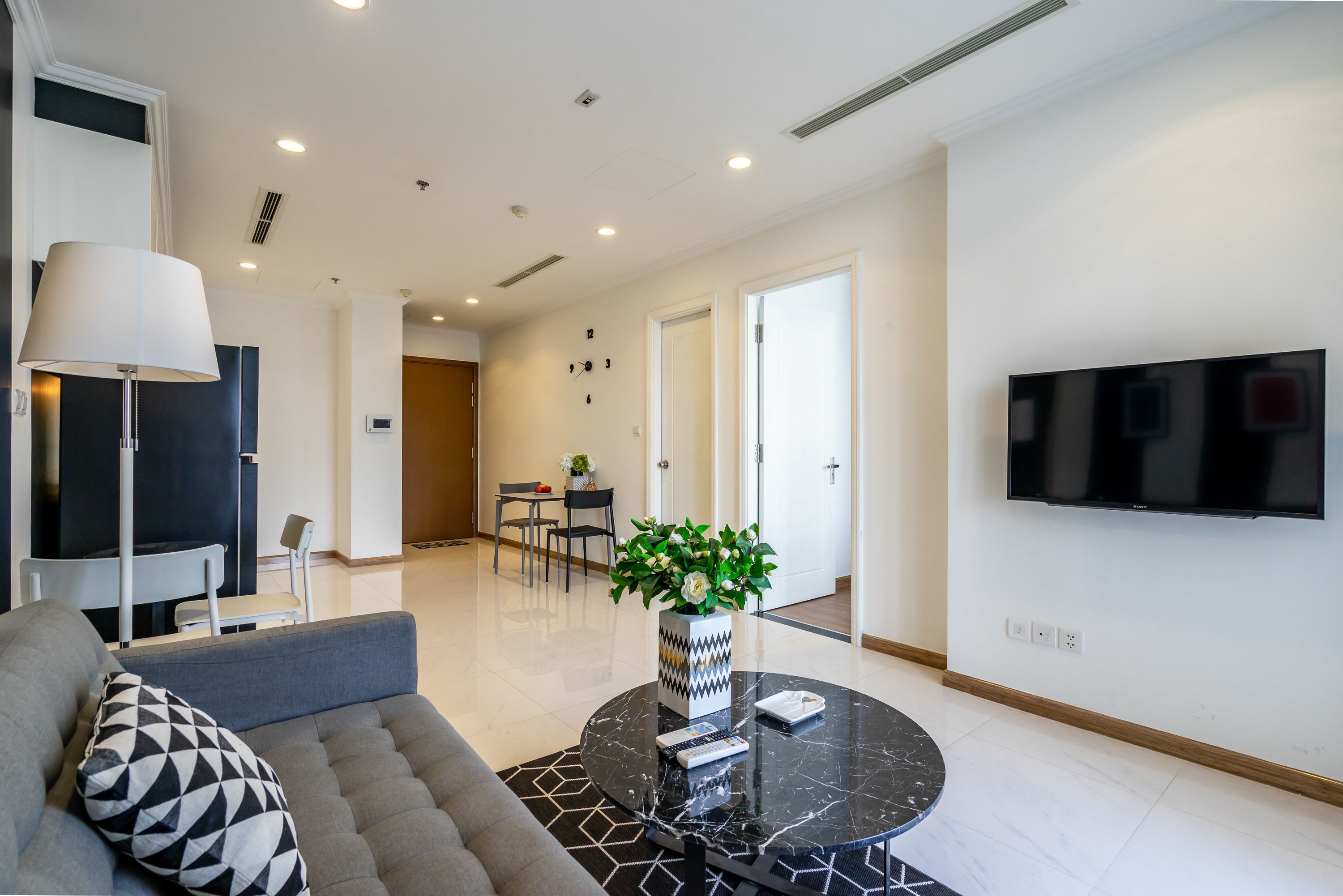A modern living room with a sofa, a round coffee table with a vase of flowers, a flat-screen TV mounted on the wall, and a dining area in the background.