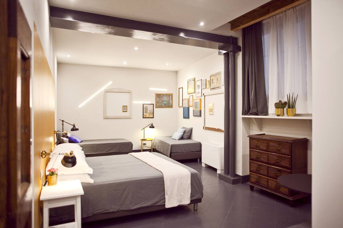 A modern hotel room with two beds, a small desk, bedside tables, a dresser, and a large window with curtains.