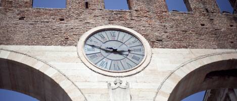 Un reloj grande montado en un arco de piedra, con una torre de ladrillo al fondo.