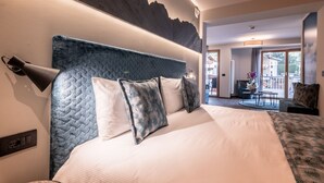 A hotel room with a large bed, a headboard with a textured design, a wall-mounted lamp, and a view of the outdoor area through a glass door. - Hotel Val di Sole (Mezzana)