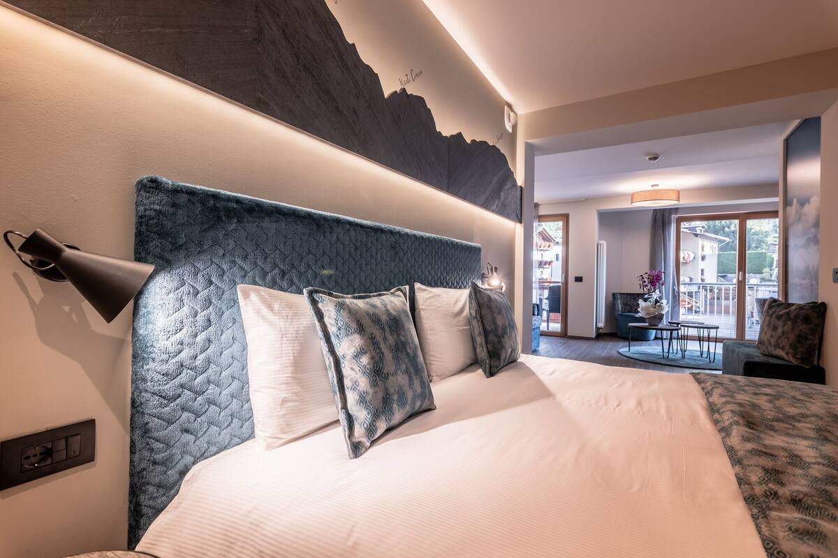 A hotel room with a large bed, a headboard with a textured design, a wall-mounted lamp, and a view of the outdoor area through a glass door.