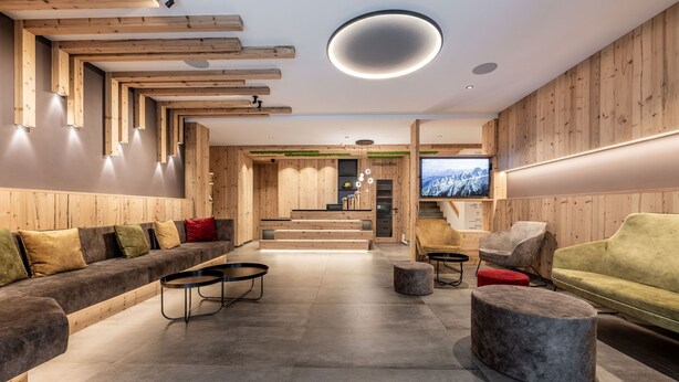 A modern interior with a wooden reception desk, a sofa, and a TV screen. - Hotel Val di Sole (Mezzana)