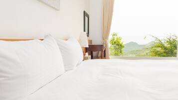 A hotel room with a large bed, a nightstand, a lamp, and a view of greenery outside.