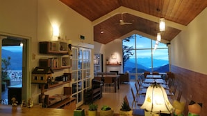 A spacious room with a high ceiling, wooden beams, and large windows offering a view of the mountains. There are bookshelves, a dining area, and a cozy seating space with potted plants. - Alishan Tea Garden (Fanlu)