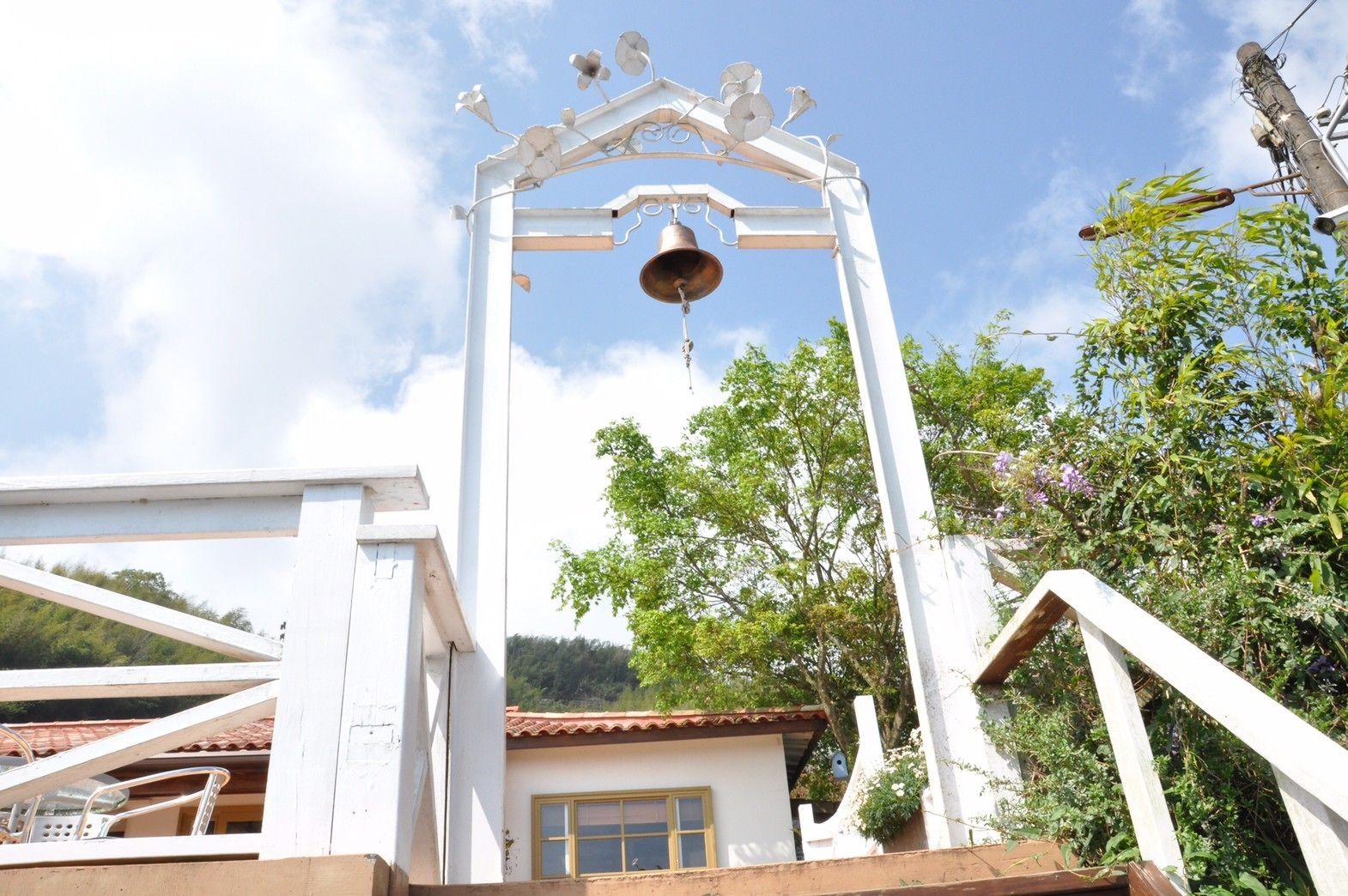 A white bell tower with a bell, surrounded by greenery and a clear sky.
