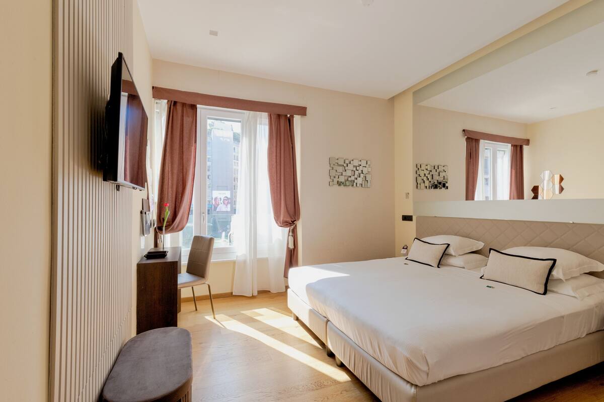A hotel room with a large bed, a desk with a chair, a television mounted on the wall, and a window with curtains.