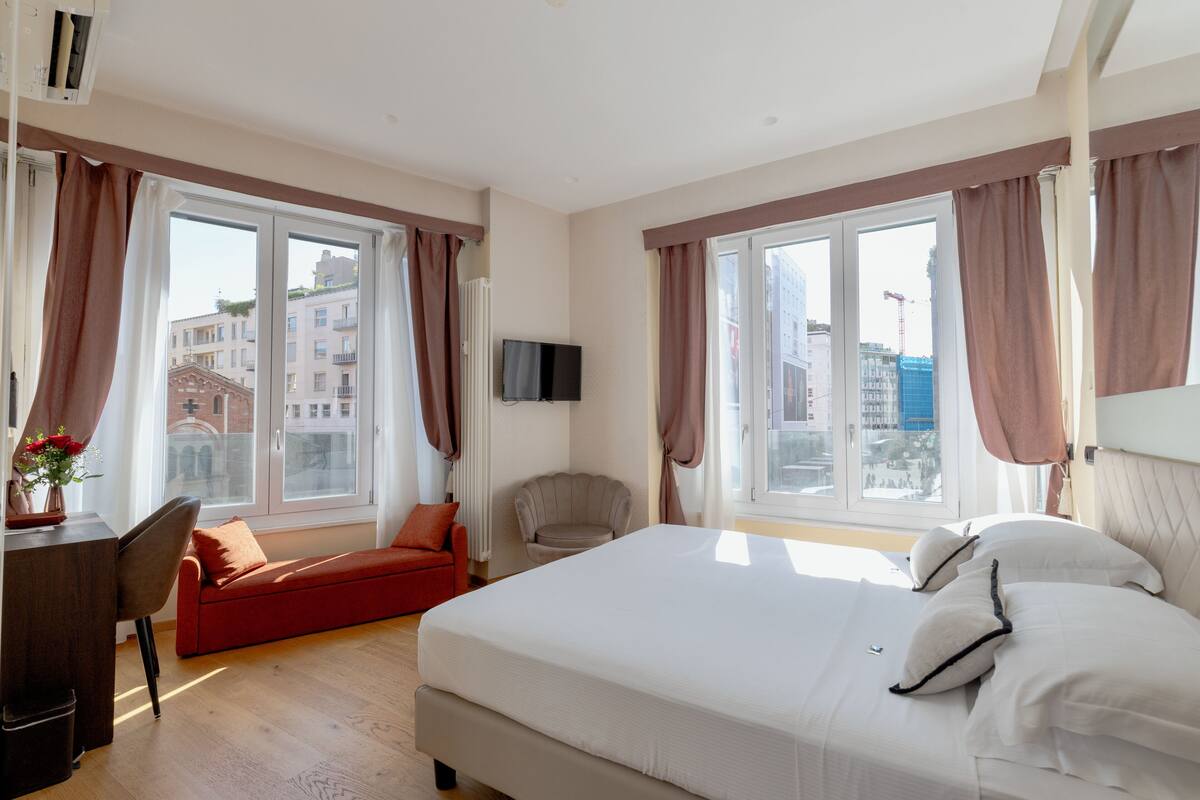 A hotel room with a large bed, a desk with a chair, a red sofa, and a TV on the wall.