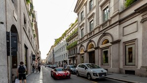 A street with historic buildings, parked cars, and pedestrians. - AH - Fashion District (Milan)