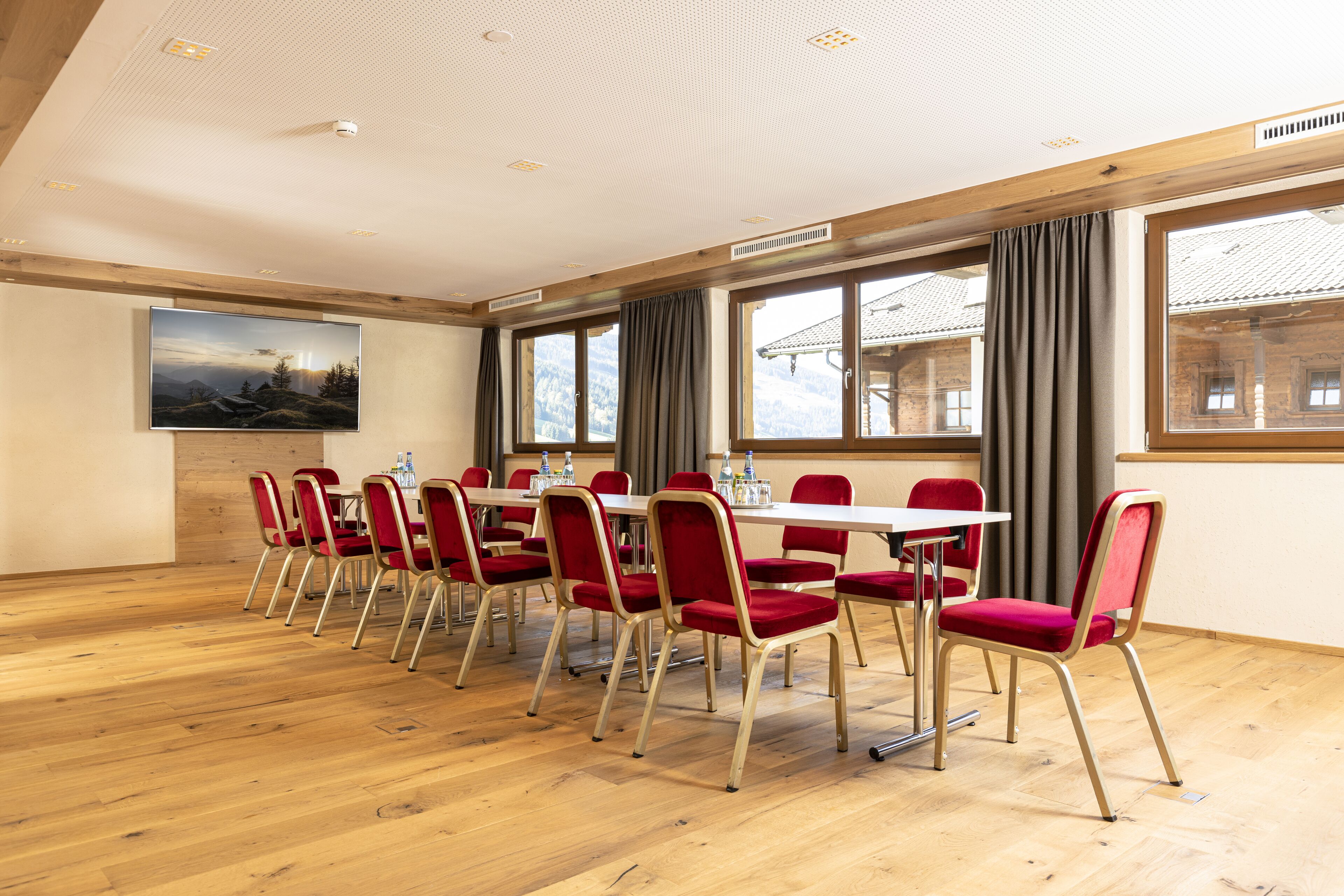 Un salón de conferencias con pisos de madera, sillas rojas, una mesa blanca y ventanas grandes con cortinas.