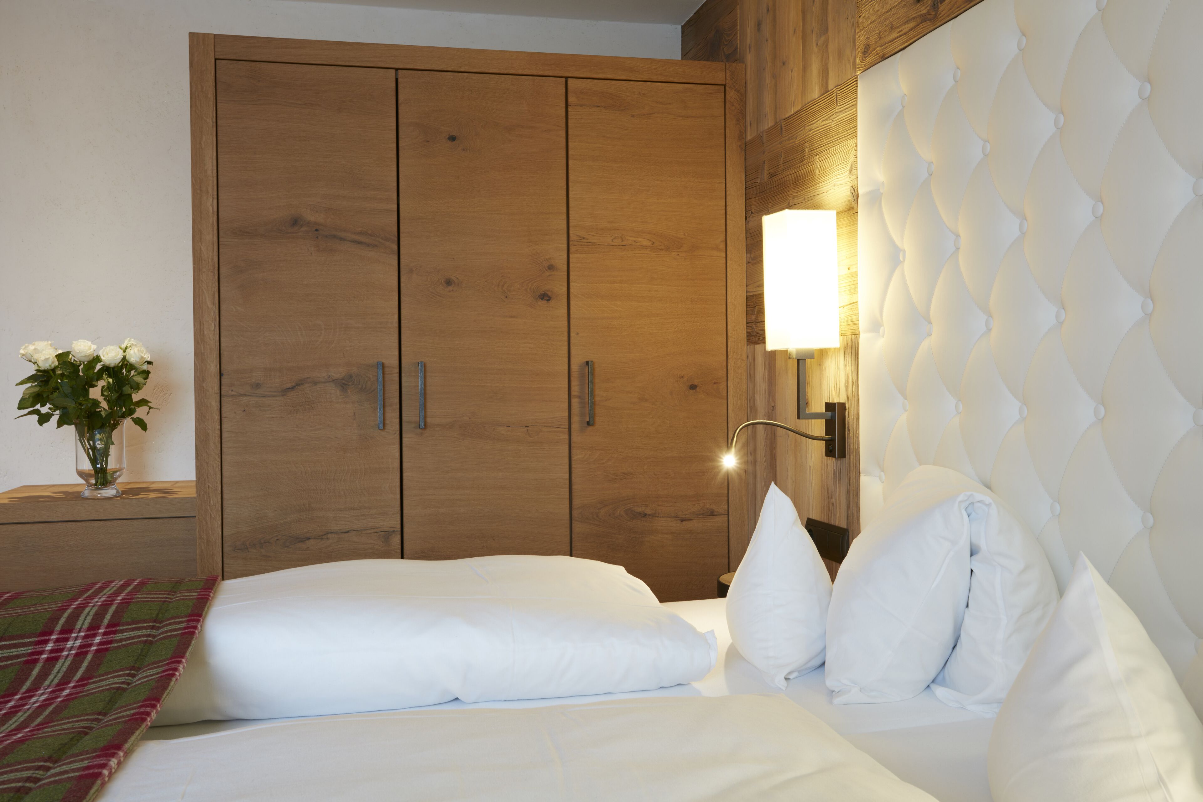 A bedroom with a wooden wardrobe, a bedside table with a lamp, and a bed with white bedding.