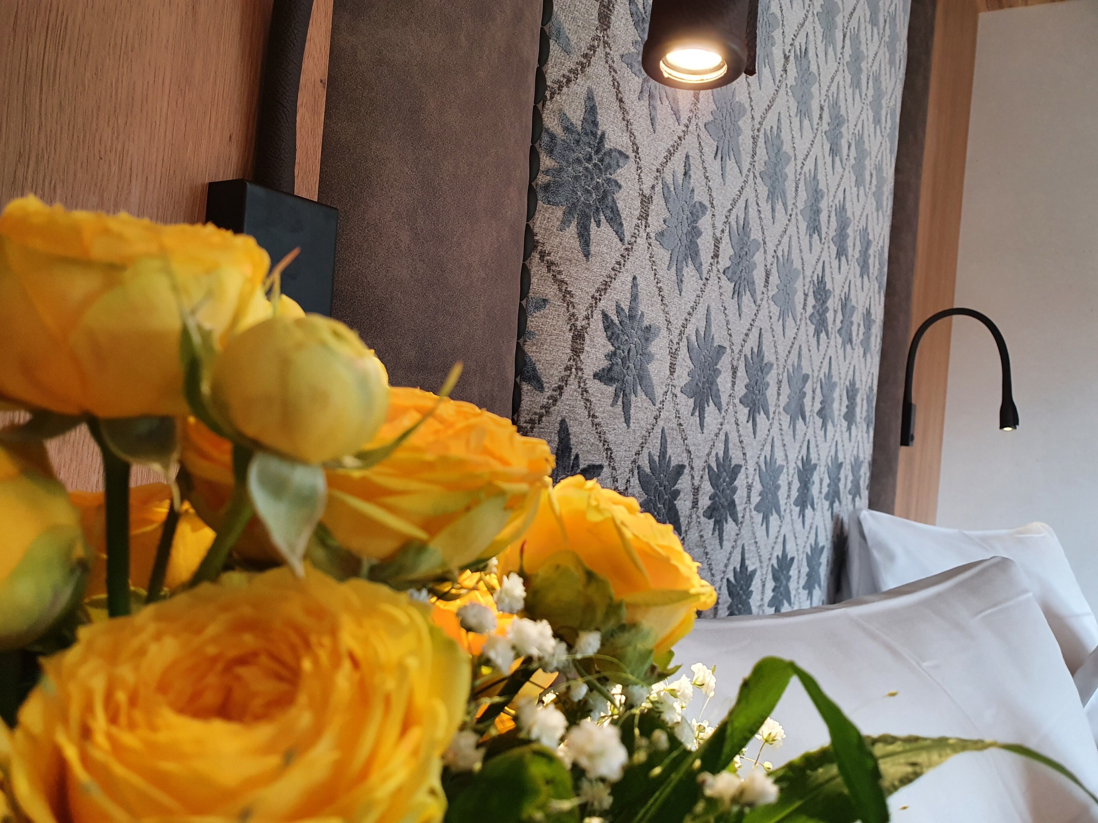 Ein Blumenstrauß aus gelben Rosen auf einem Tisch, im Hintergrund eine Wand mit Muster und eine moderne Leuchte.