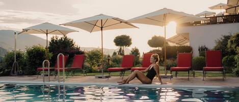 Un área junto a la piscina con sillas y sombrillas rojas, una piscina cristalina y un paisaje montañoso de fondo.