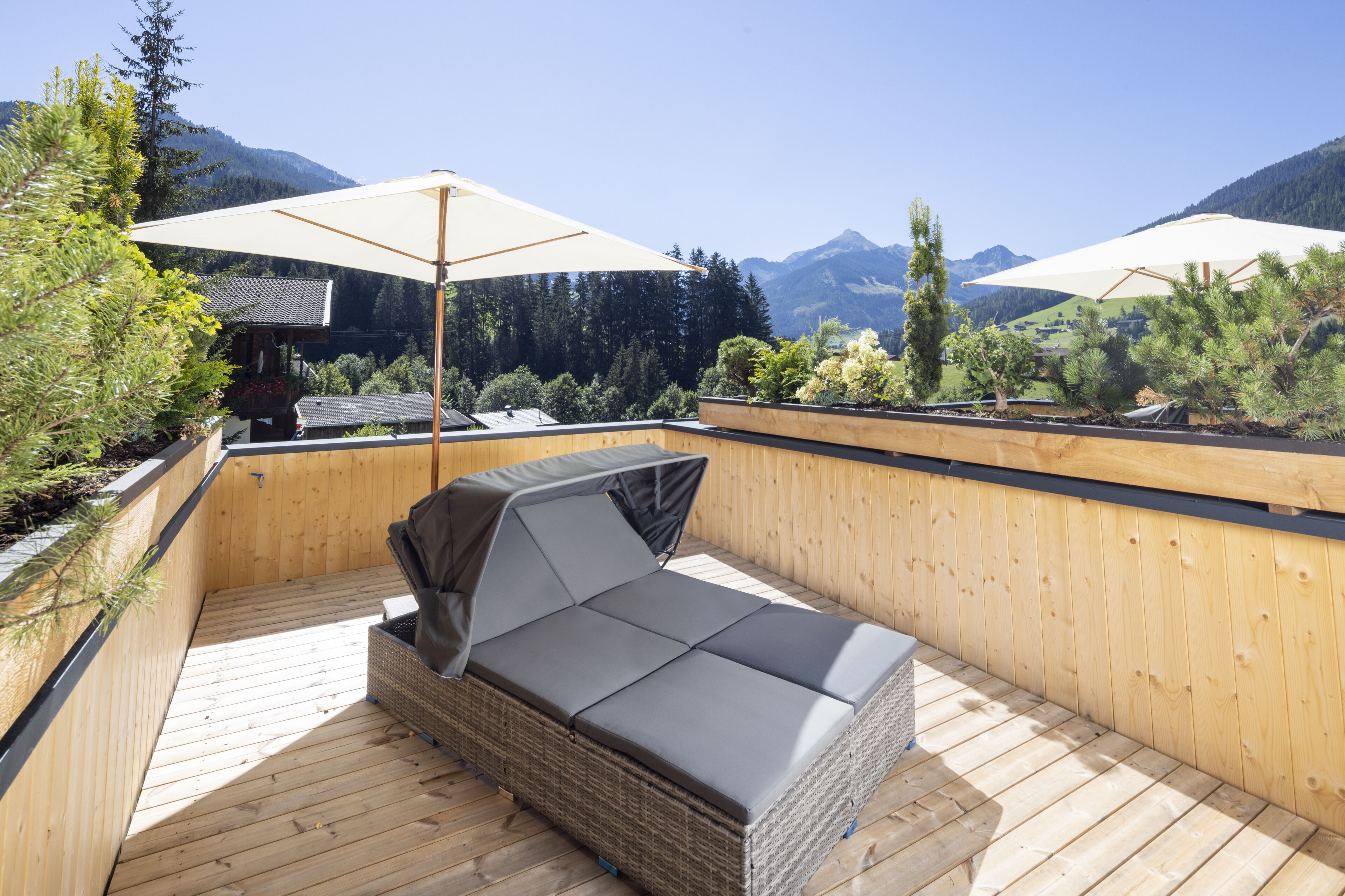 A wooden deck with a wicker sofa and two large umbrellas, overlooking a mountainous landscape.