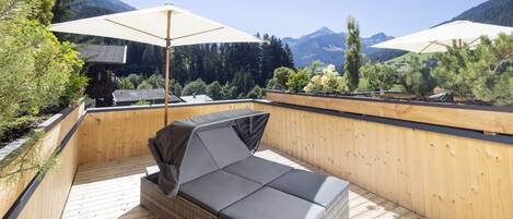 A wooden deck with a wicker sofa and two large umbrellas, overlooking a mountainous landscape.