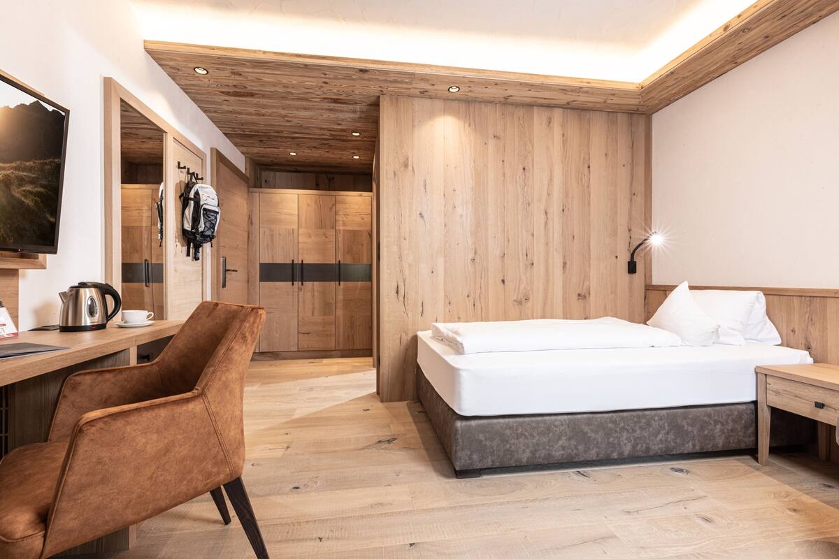 A modern hotel room with a wooden ceiling, a bed, a desk with a chair, a TV, and a wardrobe.