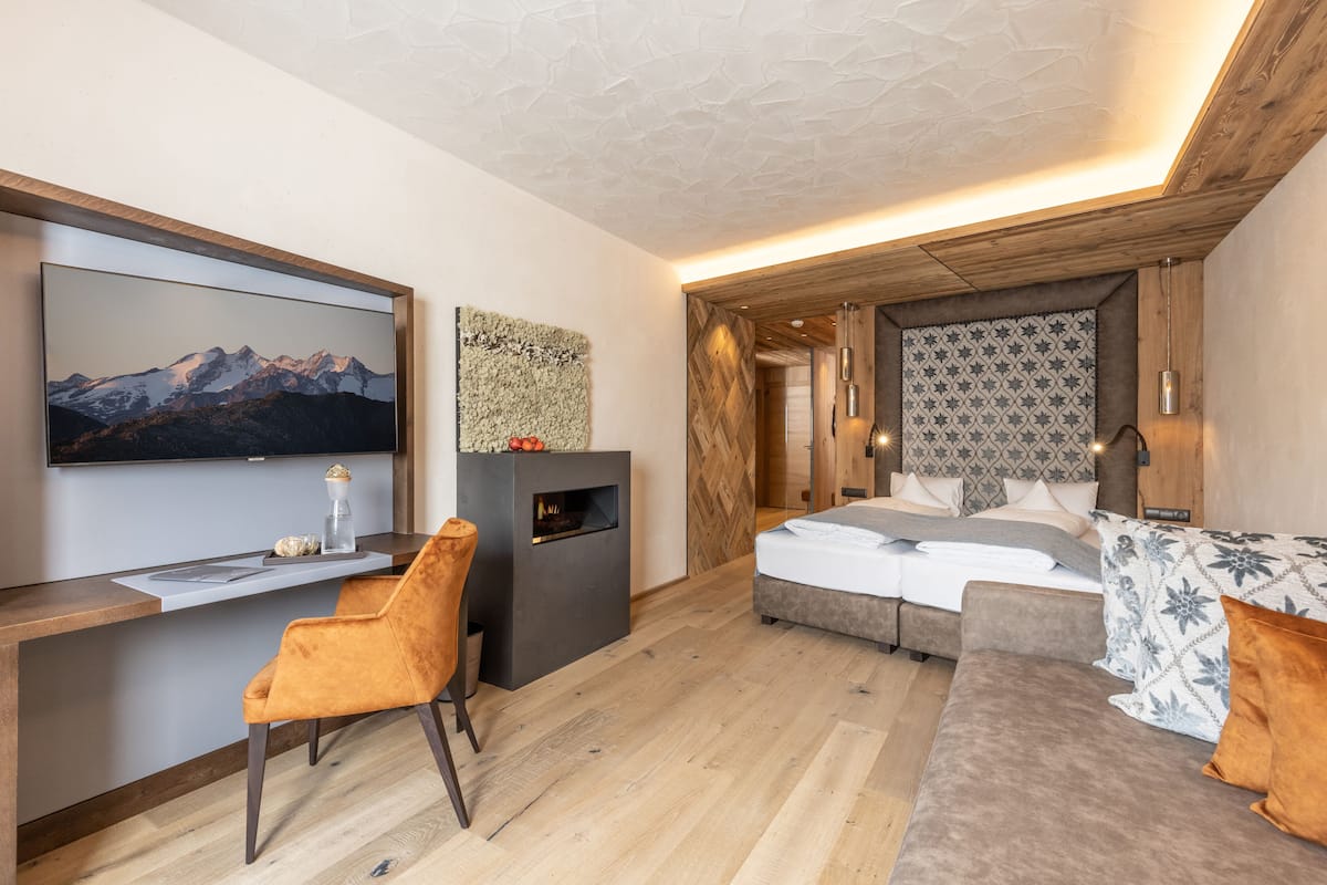 A modern hotel room with a large bed, a sofa, a desk with a chair, a fireplace, and a TV mounted on the wall.