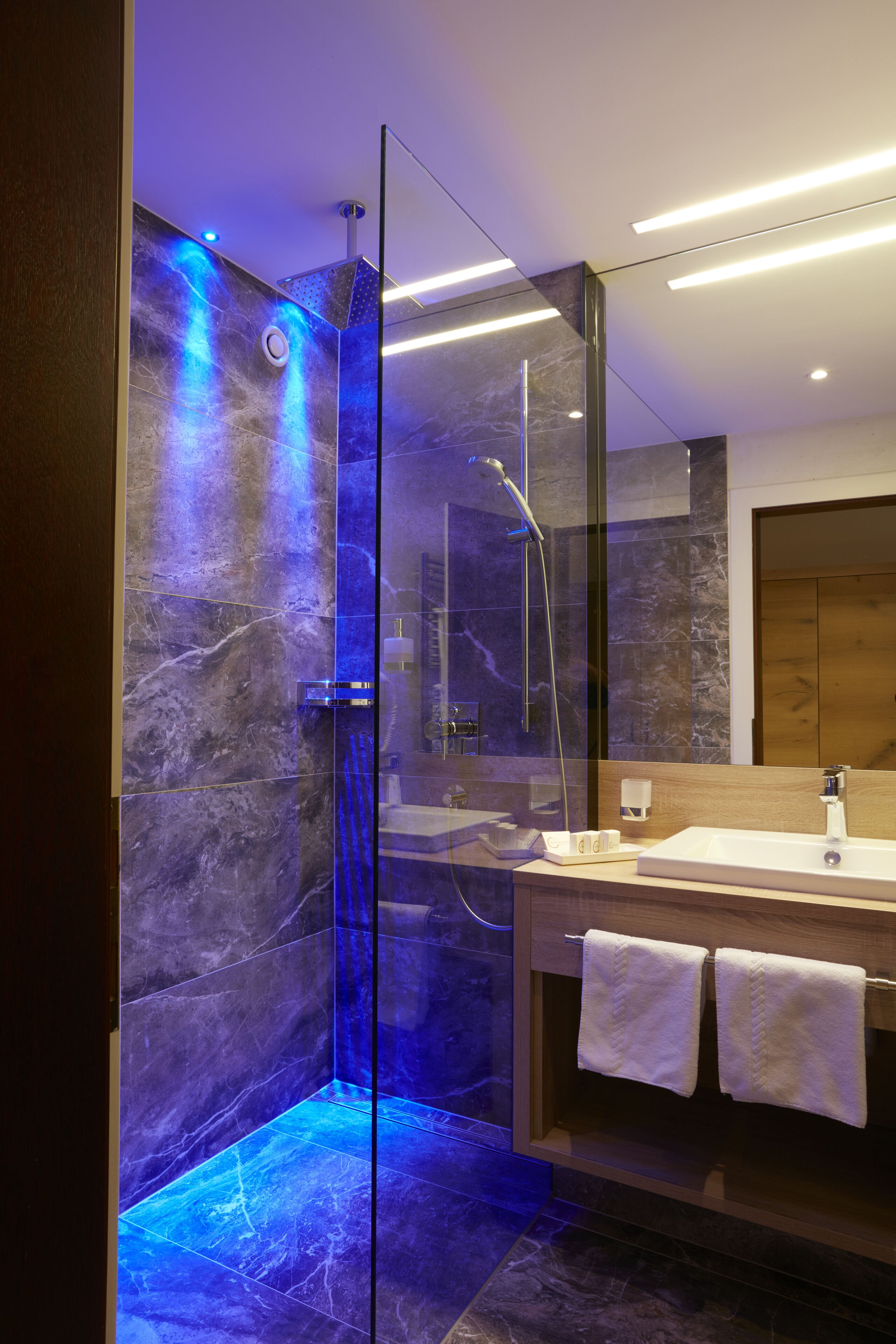 A modern bathroom with a glass-enclosed shower, marble walls, and a wooden vanity with two towels.