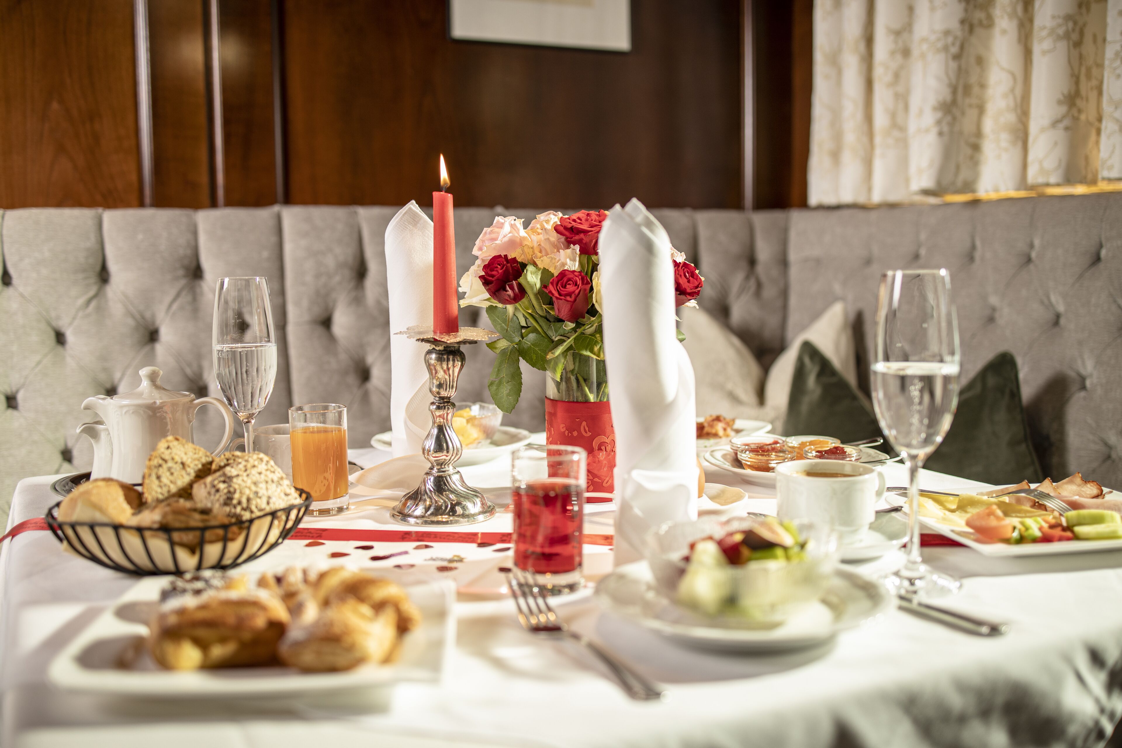 A dining table set with breakfast items, including pastries, fruit, and beverages, in a room with a tufted sofa and a vase of flowers.