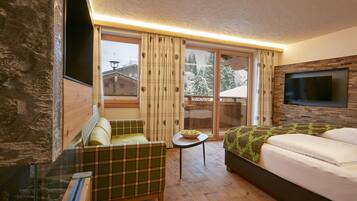 A room with a sofa, a bed, a TV mounted on the wall, and a view of snowy trees through the windows.