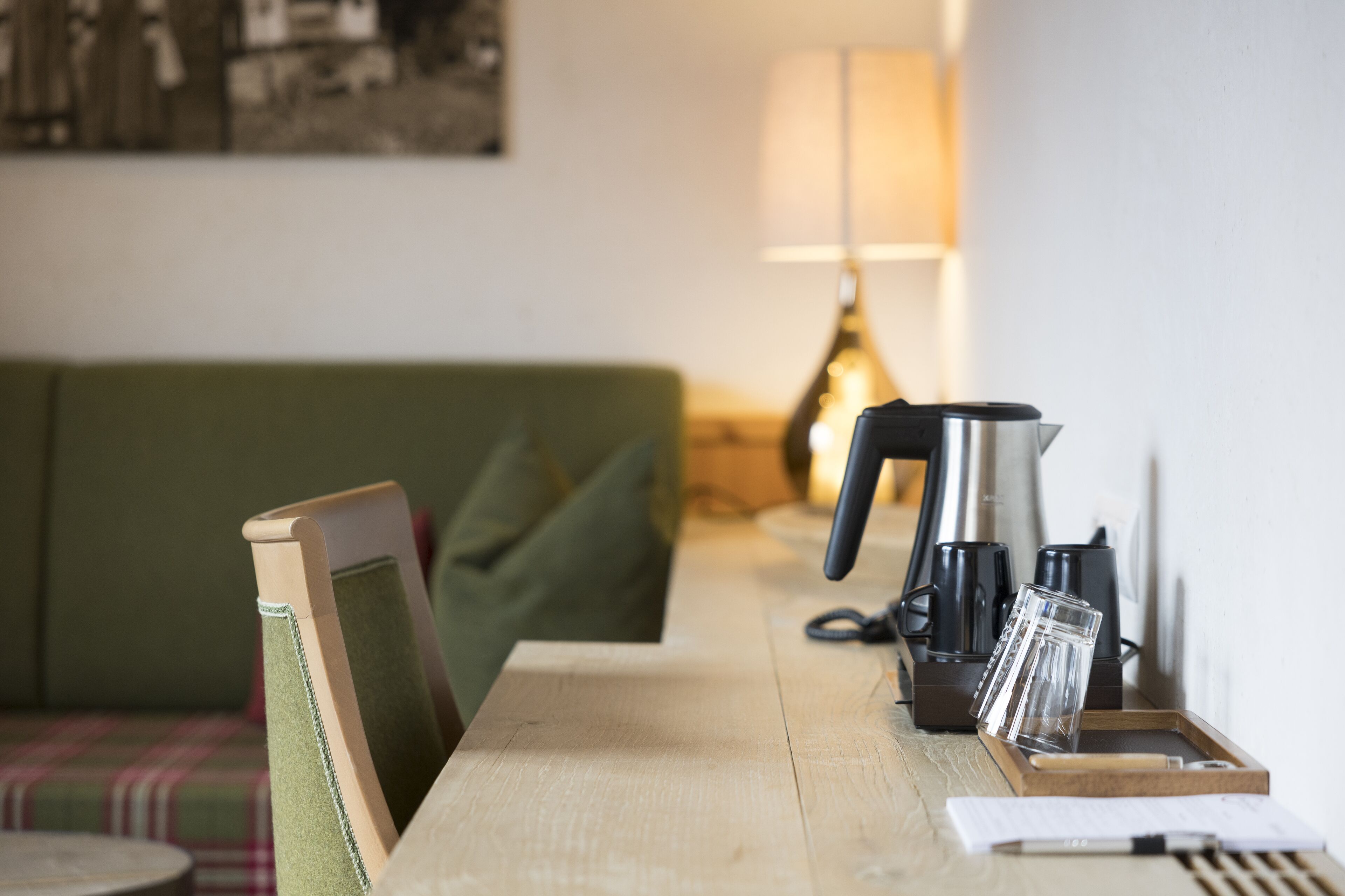A wooden table with a kettle, coffee maker, and glass. A green sofa and a lamp are in the background.