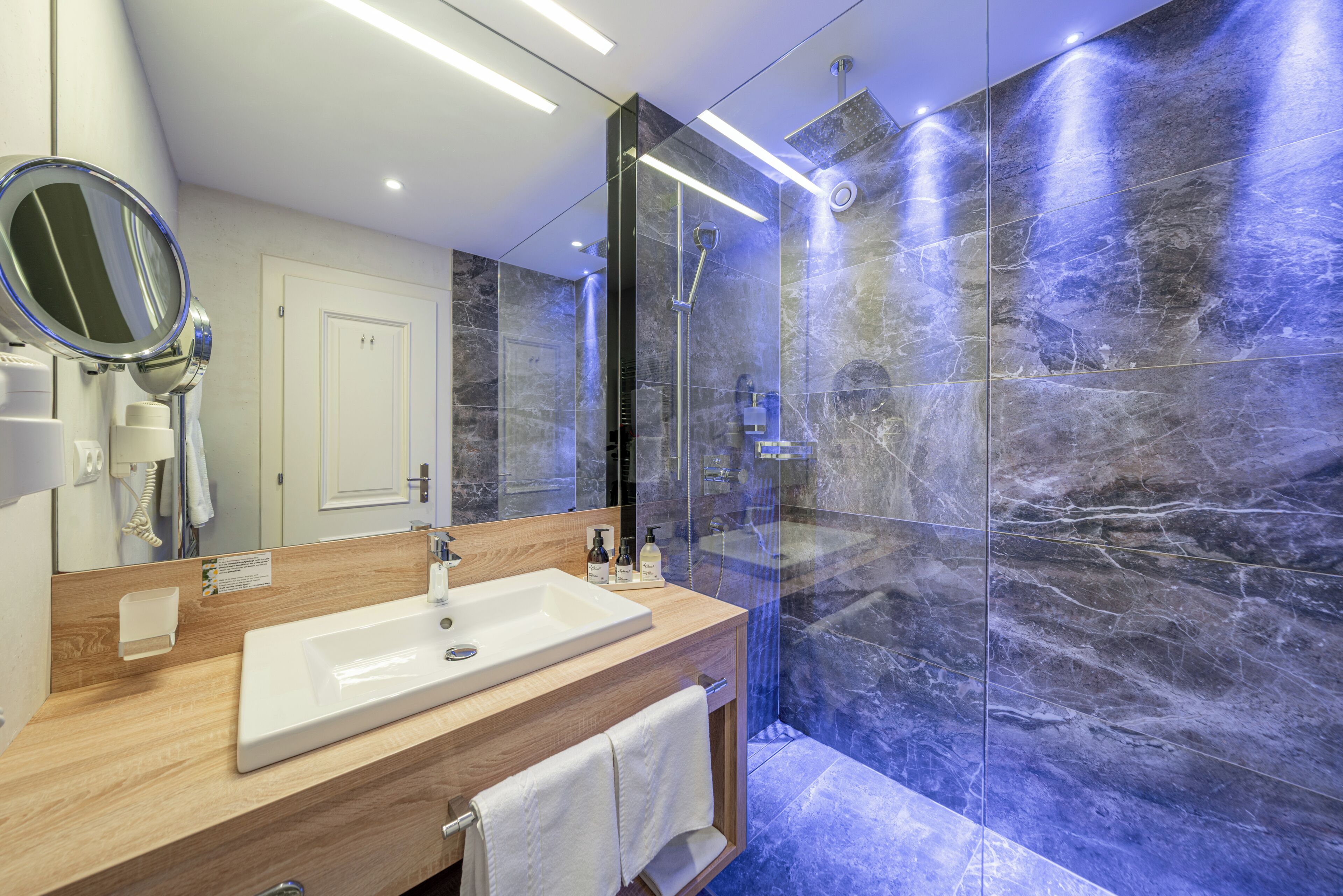 A modern bathroom with a large mirror, a sink, a shower, and marble walls.