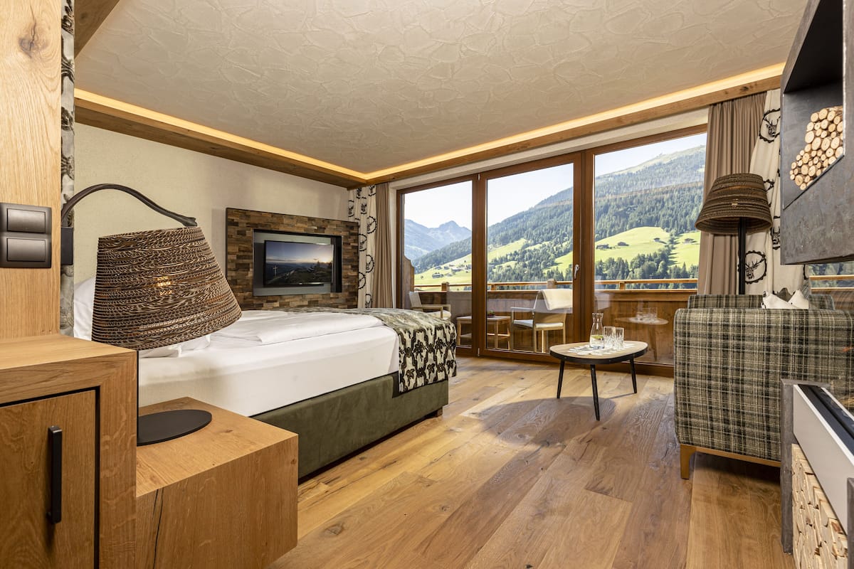A modern bedroom with a large bed, a TV, a wooden desk, and a view of mountains through large windows.