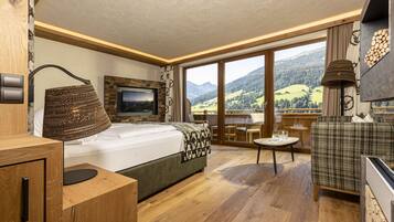 A modern bedroom with a large bed, a TV, a wooden desk, and a view of mountains through large windows.