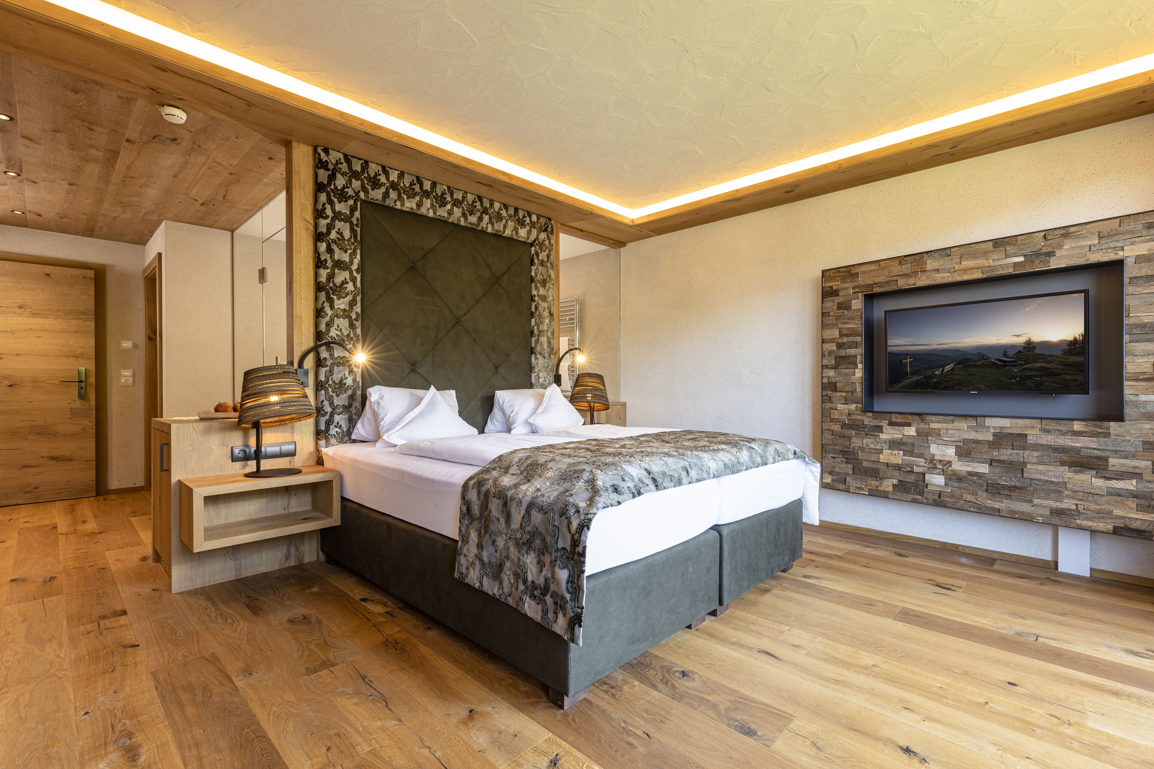 A modern bedroom with a large bed, wooden flooring, a stone accent wall, and a mounted TV.