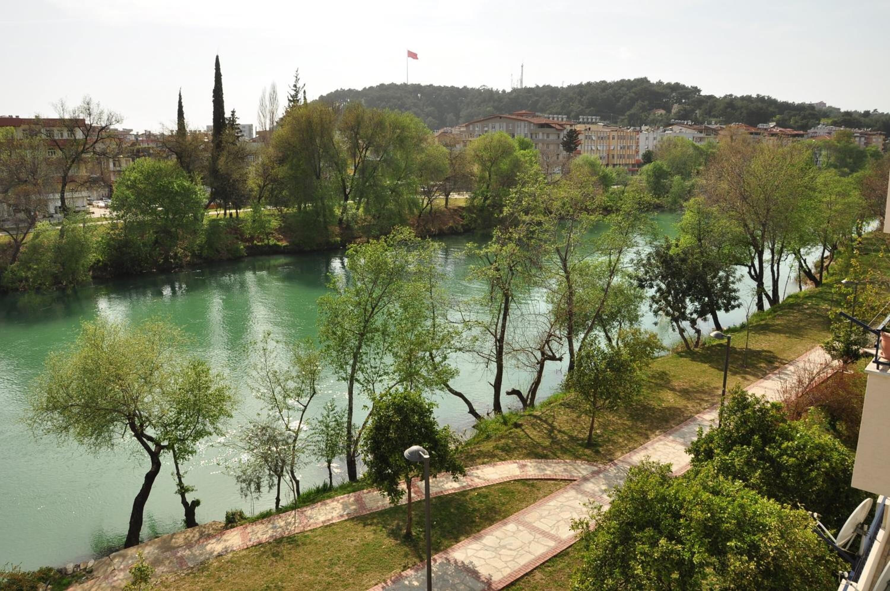 A riverside view with a pathway, trees, and buildings in the background.