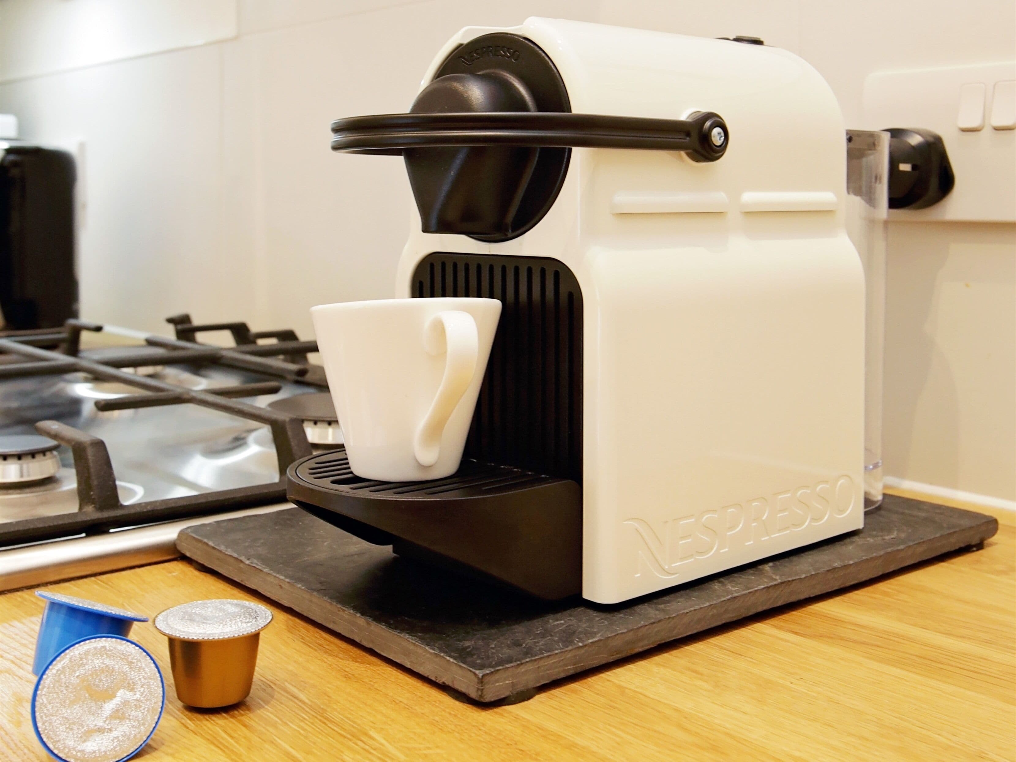 Eine Nespresso-Kaffeemaschine auf einer Küchenarbeitsplatte mit Tassenhalter, Herd und einem kleinen blauen Behälter.
