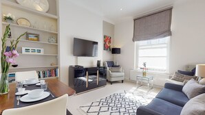 Una sala de estar moderna con un televisor de pantalla plana, una zona de comedor con una mesa dispuesta para dos personas, un sofá y un ventanal con vistas.
